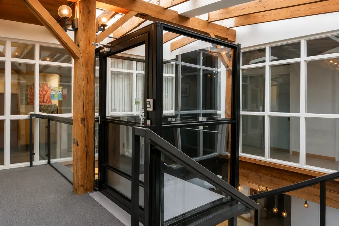 A modern glass elevator with black framing is installed in a hallway with exposed wooden beams and large windows.