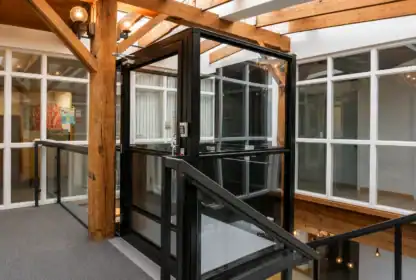 A modern glass elevator with black framing is installed in a hallway with exposed wooden beams and large windows.