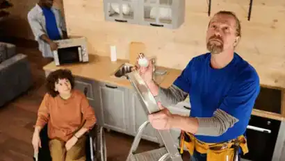 A man on a ladder changes a lightbulb in a kitchen while a woman in a wheelchair watches and another man uses a microwave in the background.