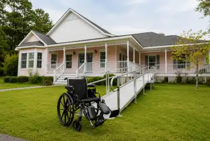 A wheelchair sits on the lawn in front of a single-story house with a white wheelchair-accessible ramp leading to the front porch.