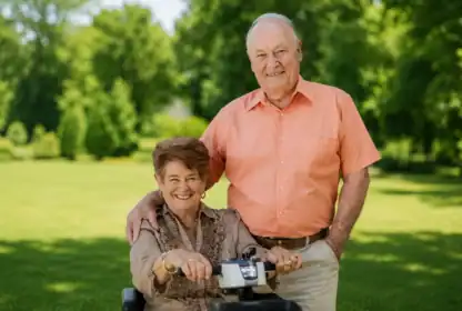 An older woman sits on a mobility scooter while an older man stands next to her outdoors in a park-like setting, both smiling at the camera.