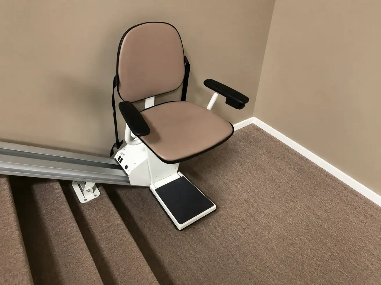 Stairlift chair installed next to a set of carpeted stairs against a beige wall, with metal rail extending up the staircase.