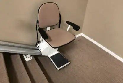 Stairlift chair installed next to a set of carpeted stairs against a beige wall, with metal rail extending up the staircase.
