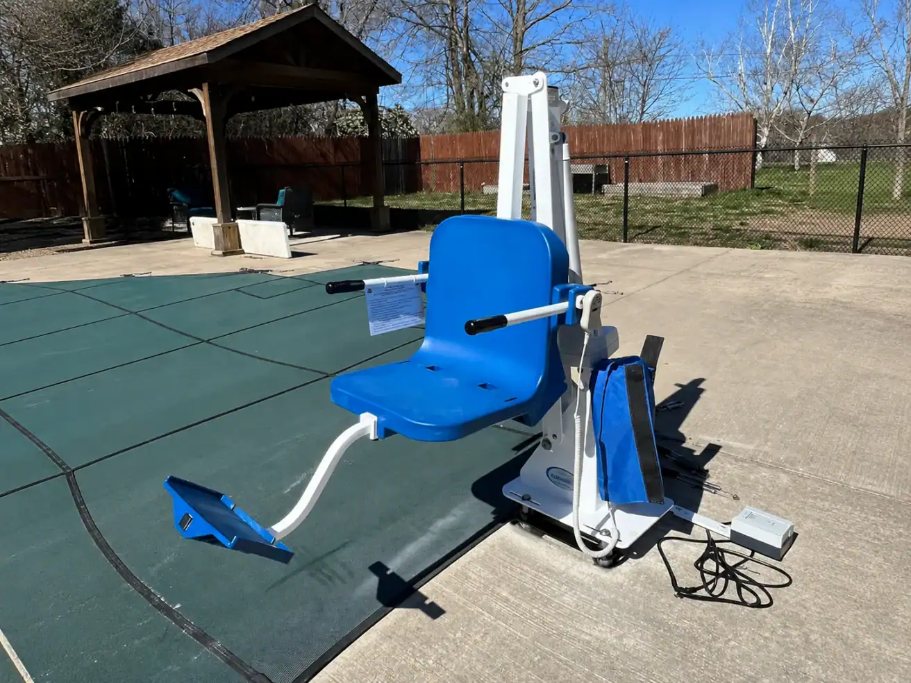 A blue and white pool lift chair is positioned next to a covered swimming pool on a sunny day, with a fenced yard and gazebo in the background.