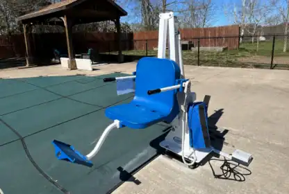 A blue and white pool lift chair is positioned next to a covered swimming pool on a sunny day, with a fenced yard and gazebo in the background.