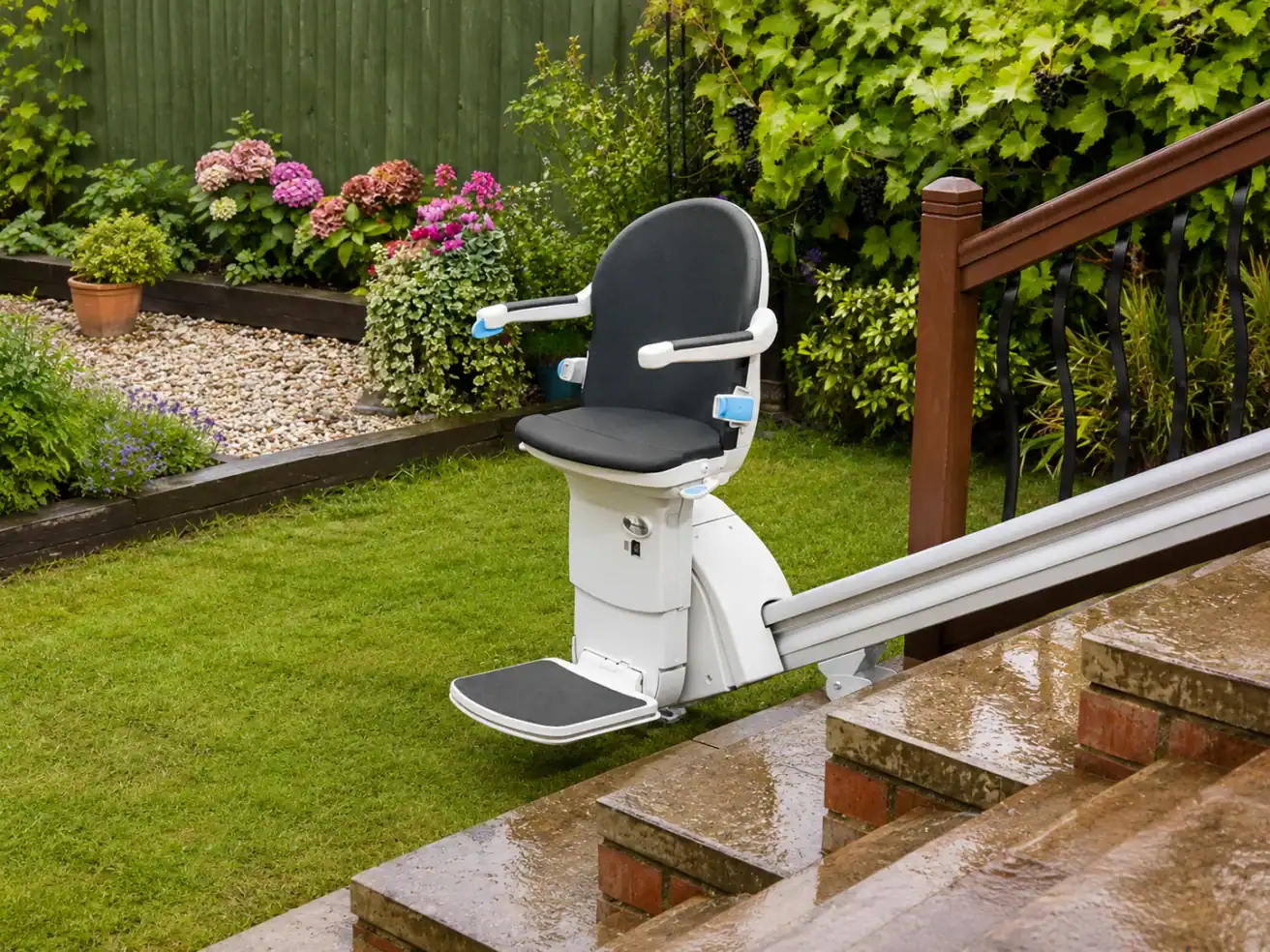 Outdoor stairlift installed on a garden staircase, next to a wet lawn and flower beds, on a rainy day.