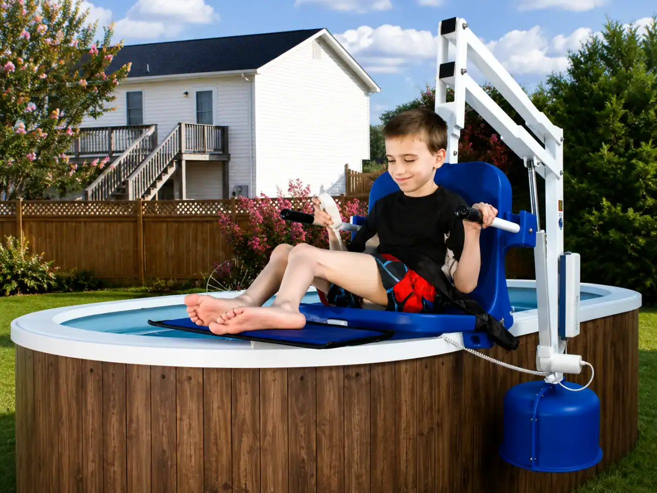 A boy sits on a pool lift chair, being lowered into an above-ground backyard pool, with a house and trees in the background.