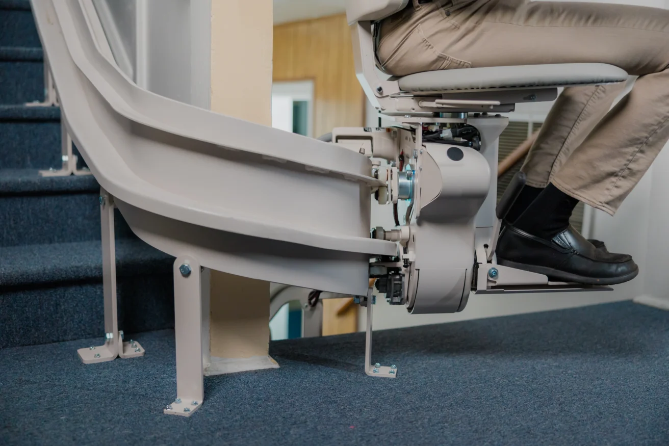 A person sits on a stair lift attached to a staircase, showing the mechanical components and tracks on a carpeted floor.