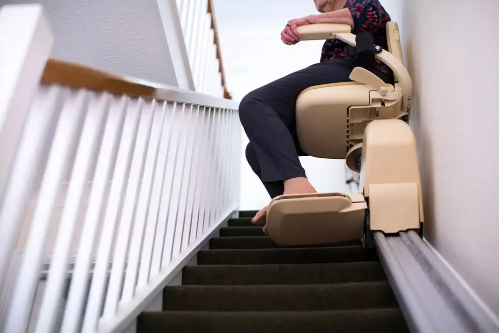 A person sits on a stairlift at the top of a carpeted staircase in a residential setting.