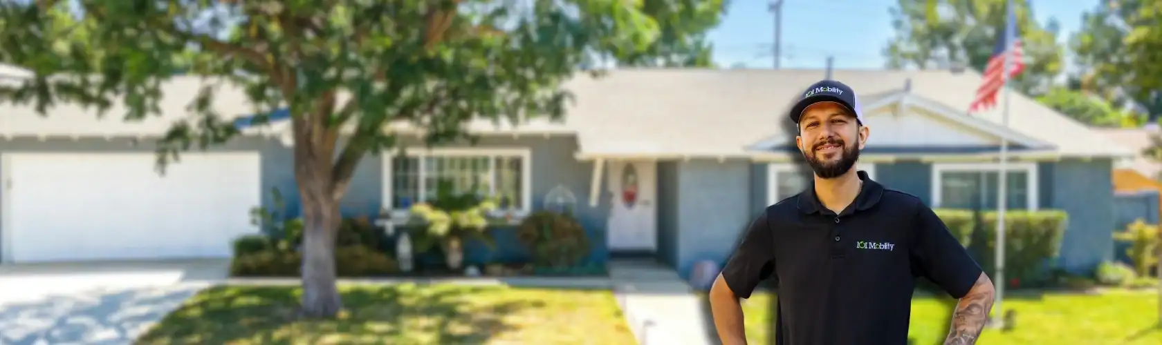 A man in a black polo shirt and cap stands smiling in front of a suburban house with a lawn, tree, and an American flag.