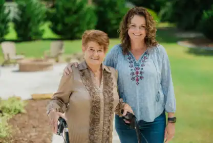 Two women stand outdoors, one older with a walker and one younger with her arm around the other, both smiling at the camera.