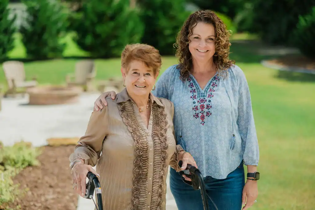 Two women stand outdoors, one older with a walker and one younger with her arm around the other, both smiling at the camera.