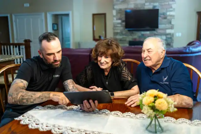 A man shows a tablet to an older woman and man sitting at a dining table with a lace tablecloth and a flower arrangement.