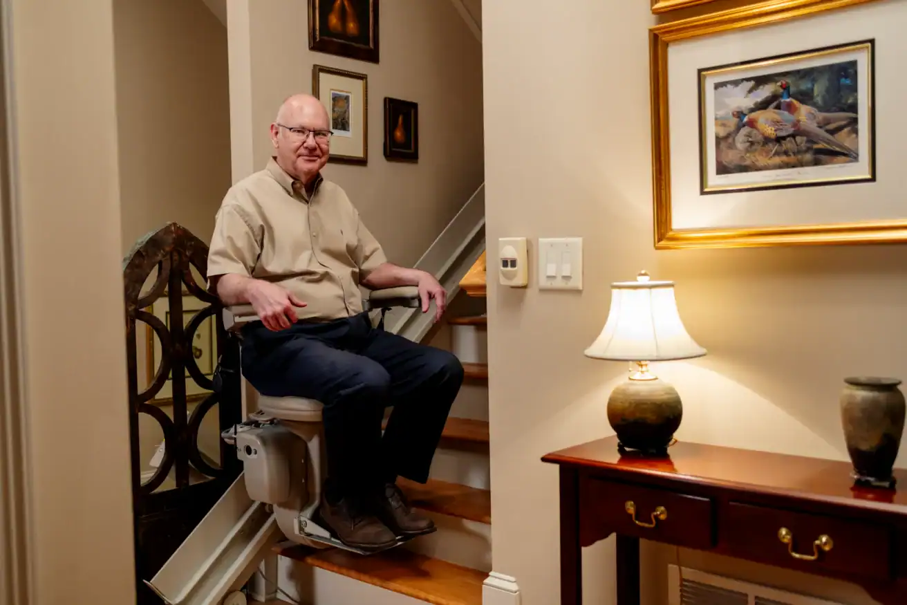 An older man sits on a stairlift at the bottom of a staircase in a well-decorated home, smiling at the camera.