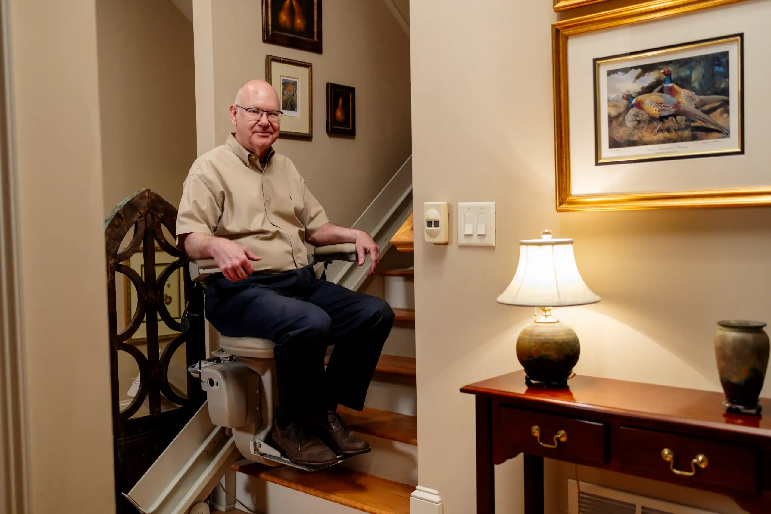 An older man sits on a stairlift at the bottom of a staircase in a well-decorated home, smiling at the camera.