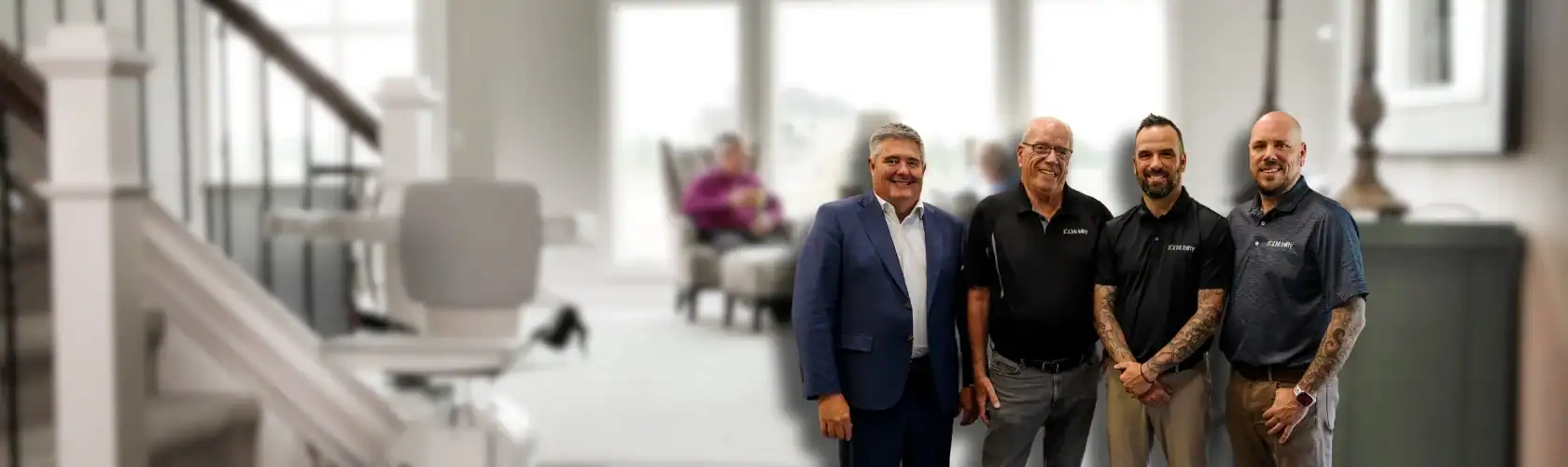 Four men stand posing in a modern, well-lit living room with a stairlift visible on the left and another person seated in the background.