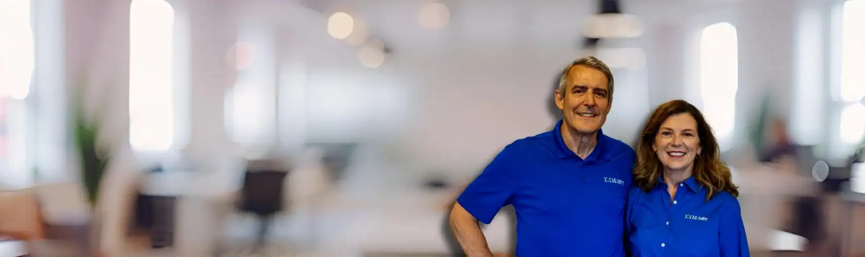 Two people wearing matching blue shirts stand together, smiling, in a brightly lit office environment with blurred background.
