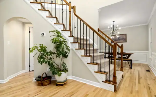 Carpeted staircase with wooden handrail, black spindles, and decorative trim in a home. Potted plant and basket at the base; dining area with chandelier visible in the background.