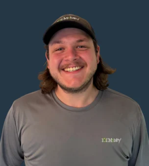 A man with long hair and a beard, wearing a gray shirt and cap with "101 Mobility" logos, smiles in front of a plain dark background.