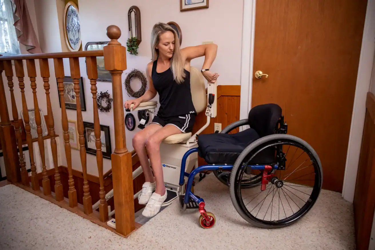 A woman transfers from a blue wheelchair to a stair lift chair at the top of a staircase inside a home.