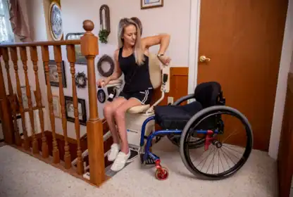 A woman transfers from a blue wheelchair to a stair lift chair at the top of a staircase inside a home.