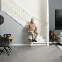 An older man sits on a stairlift attached to a white staircase in a modern, open-plan living and dining room.