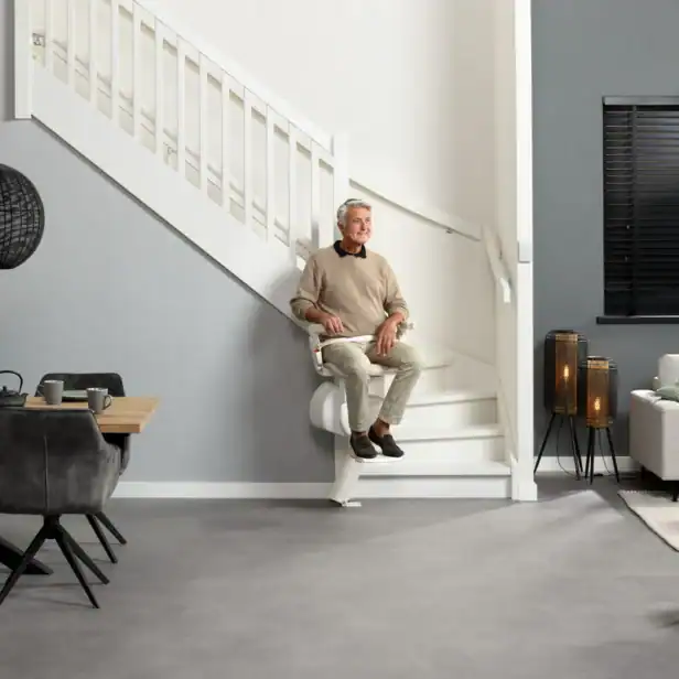 An older man sits on a stairlift attached to a white staircase in a modern, open-plan living and dining room.