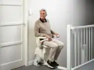 An older man sits on a stair lift chair at the top of a staircase in a modern indoor setting, facing sideways and looking toward the door.