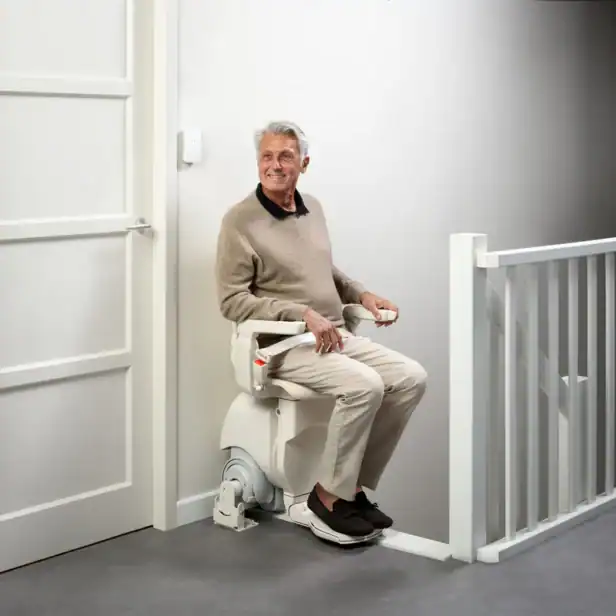 An older man sits on a stair lift chair at the top of a staircase in a modern indoor setting, facing sideways and looking toward the door.