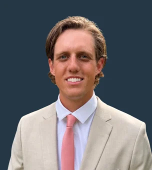 Man in a beige suit with a white shirt and pink tie standing in front of a plain dark background, smiling at the camera.