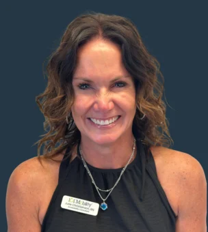 A woman with wavy brown hair wearing a sleeveless black top and a name tag smiles at the camera against a plain dark background.