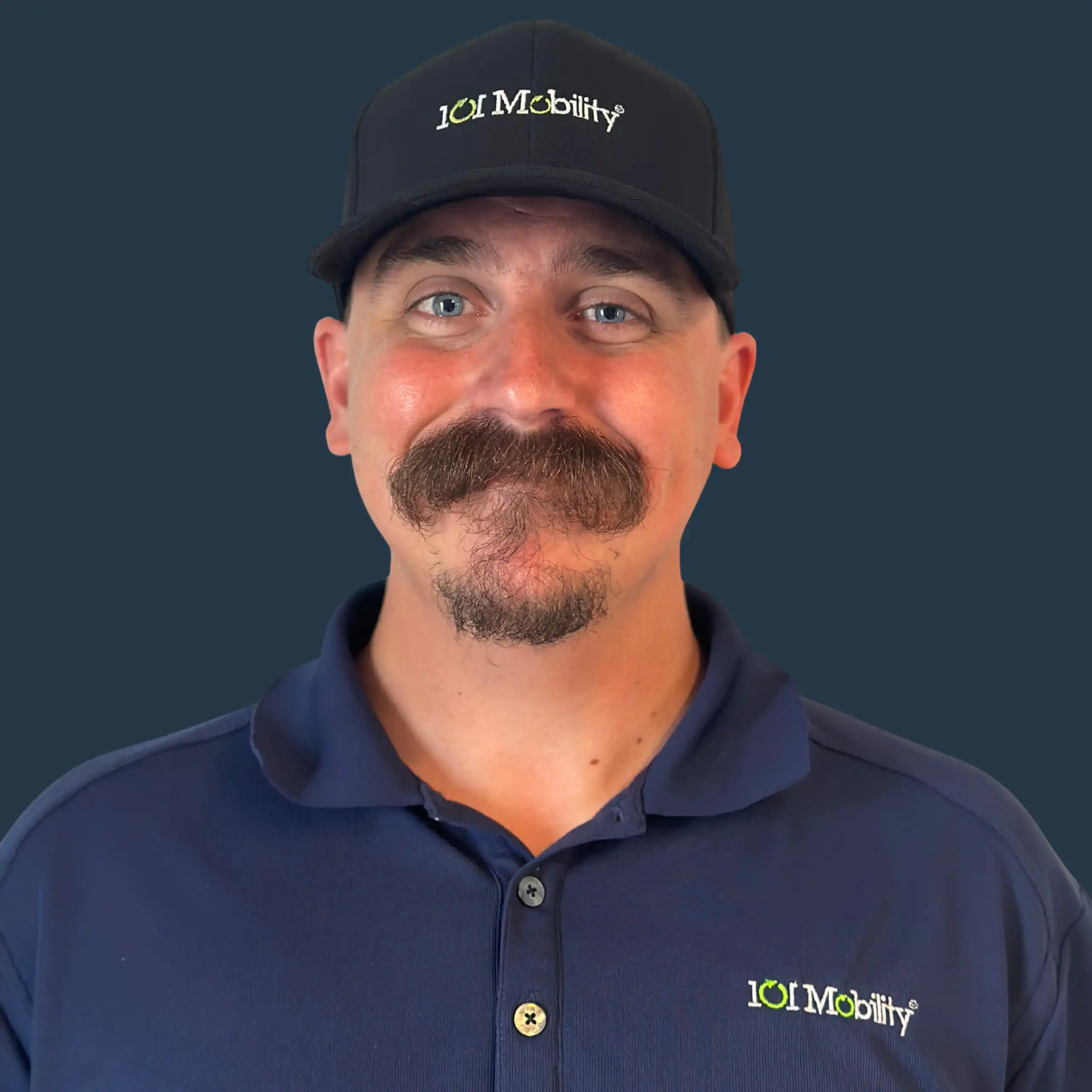 Man wearing a navy blue shirt and hat with "101 Mobility" logos, standing against a plain dark background, smiling at the camera.