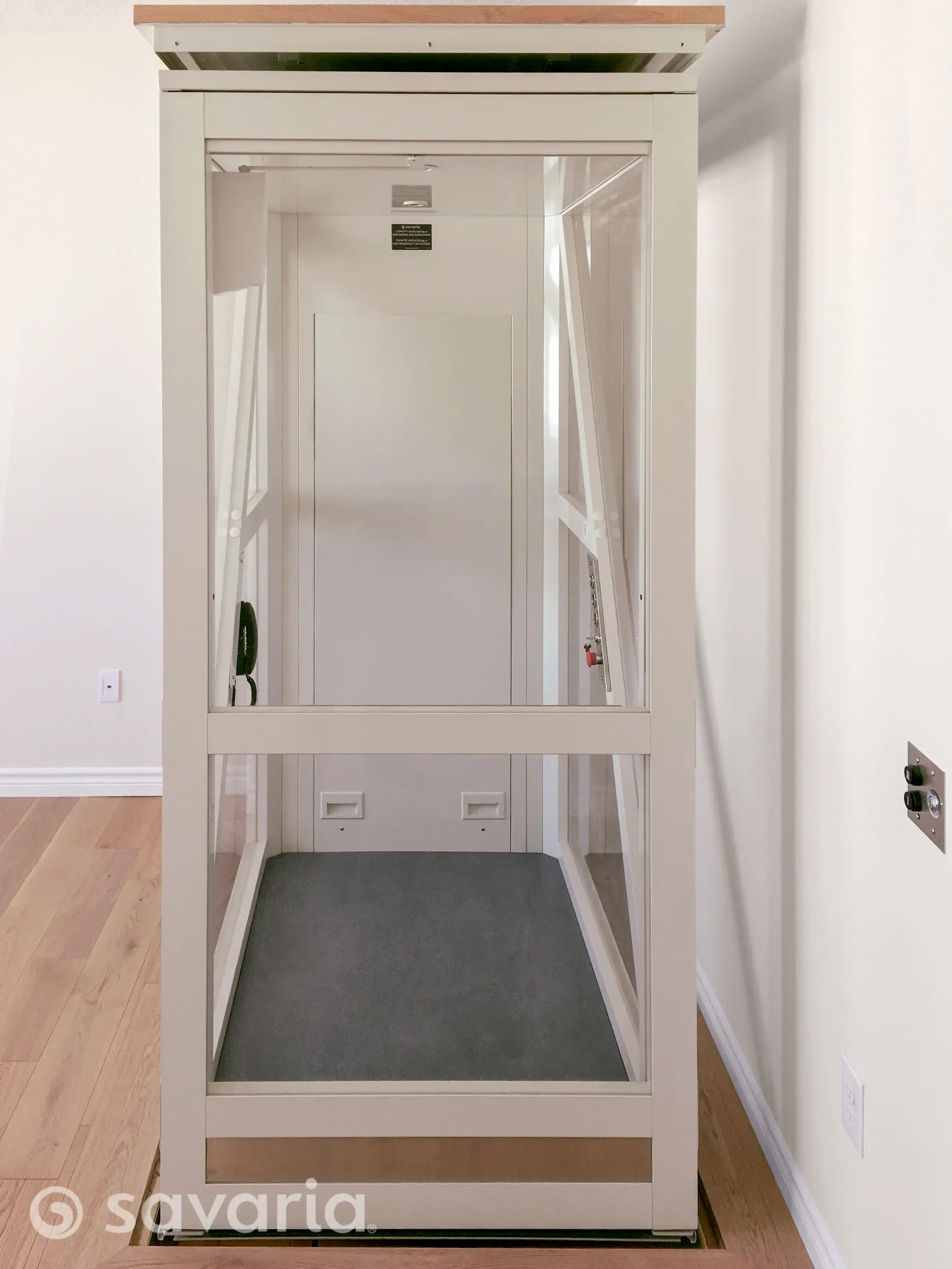 A residential home elevator with glass walls and a carpeted floor, positioned next to a white wall and a wood floor. The Savaria brand logo is visible in the lower left corner.