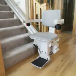 A stair lift with a gray seat is installed beside a carpeted staircase in a home with wooden floors and large windows.
