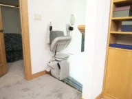 A stair lift chair is installed at the top of a staircase in a home, next to a wooden bookshelf and an open bedroom door.