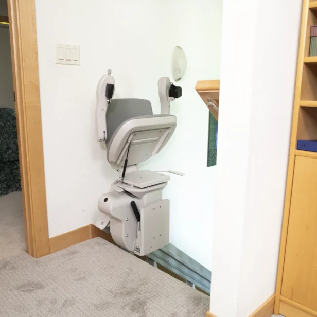 A stair lift chair is installed at the top of a staircase in a home, next to a wooden bookshelf and an open bedroom door.
