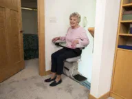 Older woman in a pink sweater sits on a stair lift chair at home, positioned near a doorway and carpeted floor.