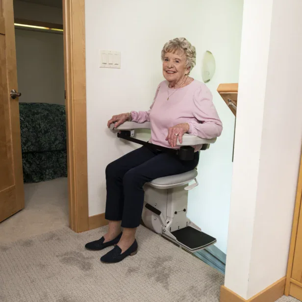 Older woman in a pink sweater sits on a stair lift chair at home, positioned near a doorway and carpeted floor.