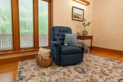 A blue recliner with pillows and a throw blanket sits on a patterned rug near large windows and a wooden side table with a vase and framed art.