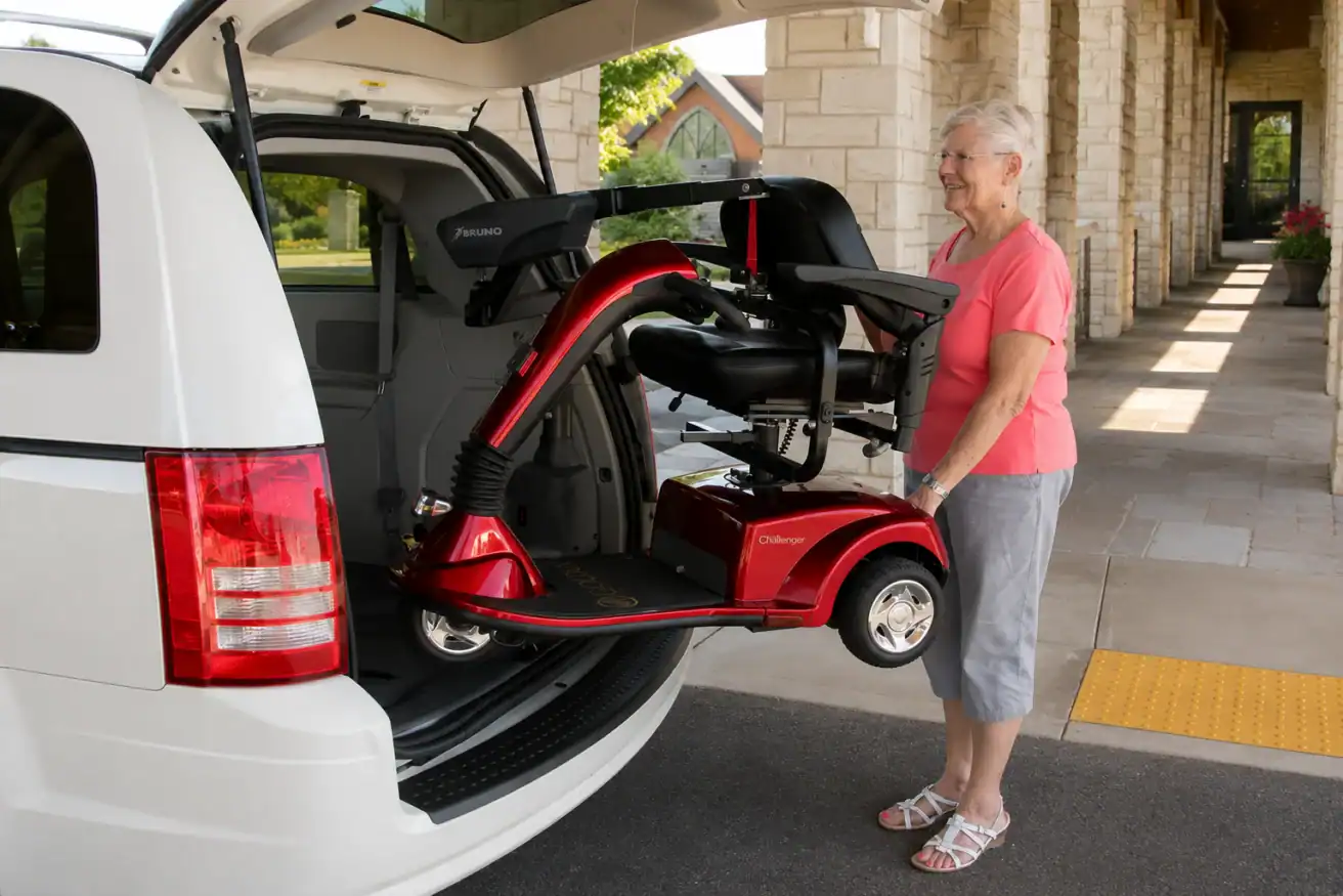 An older woman loads a red mobility scooter into the back of a white van using a lift, outside a building on a sunny day.