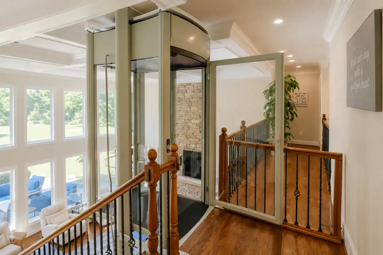 A modern glass residential elevator with an open door is installed next to a staircase in a bright home with large windows and wood flooring.