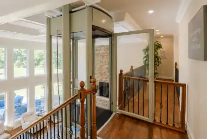 A modern glass residential elevator with an open door is installed next to a staircase in a bright home with large windows and wood flooring.