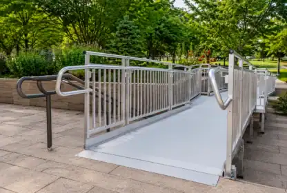A metal wheelchair ramp with handrails on both sides is installed on a paved walkway, surrounded by landscaped greenery and trees in an outdoor park setting.