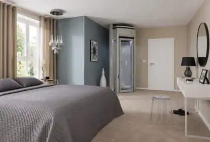 Modern bedroom with gray bedding, large window, glass shower cabin in the corner, desk with lamp, round mirror, and neutral-colored walls and carpet.