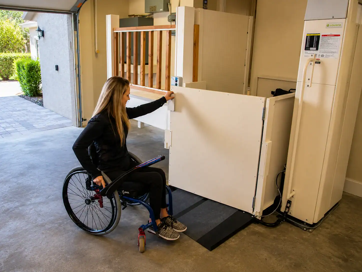 A woman in a wheelchair uses a residential vertical platform lift in a garage, raising the gate to access an elevated indoor entryway.