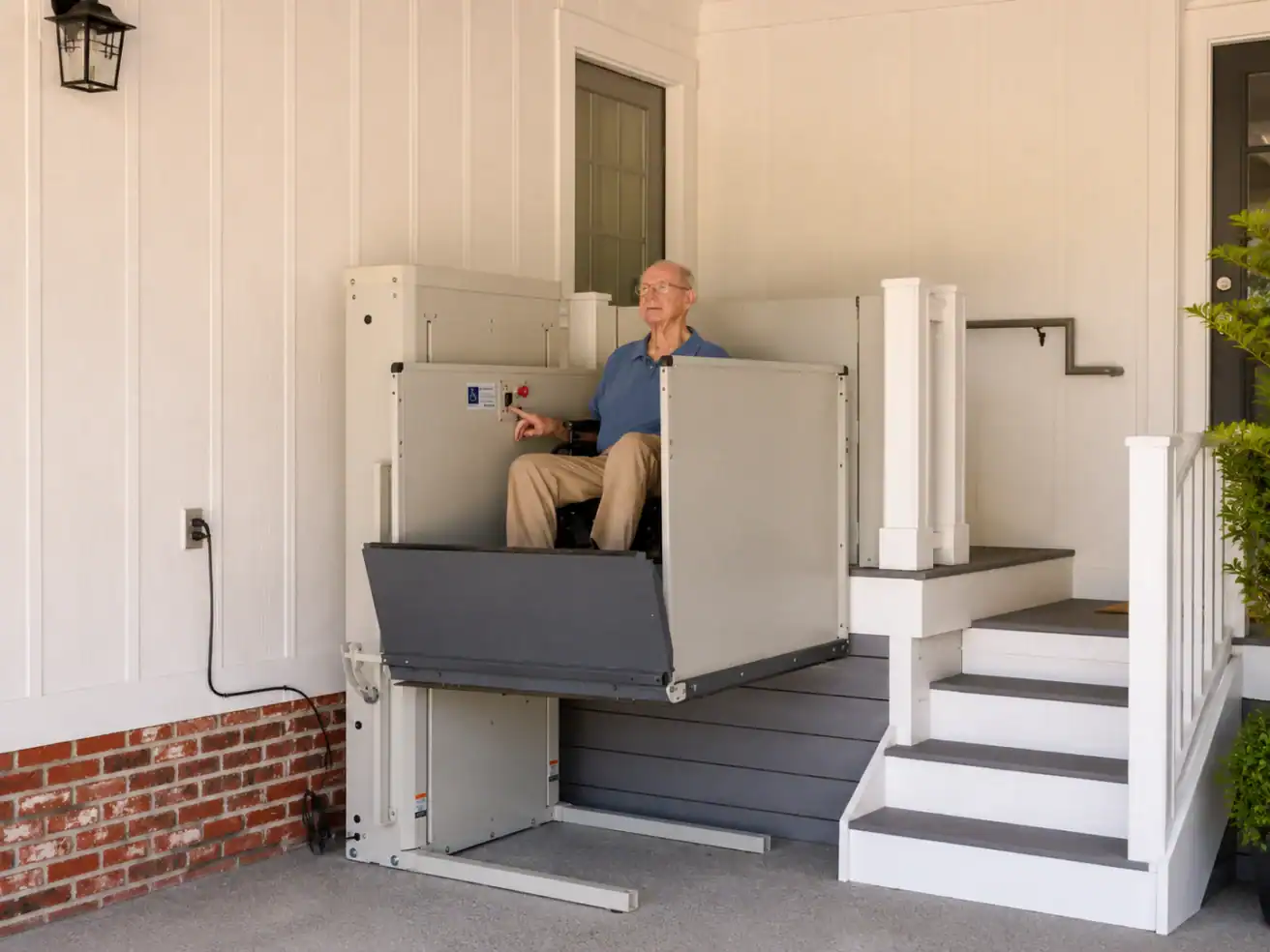 An older man in a wheelchair uses a vertical platform lift to access a porch, next to stairs leading to a door of a house.