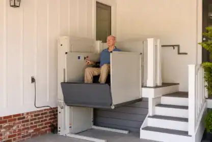 An older man in a wheelchair uses a vertical platform lift to access a porch, next to stairs leading to a door of a house.