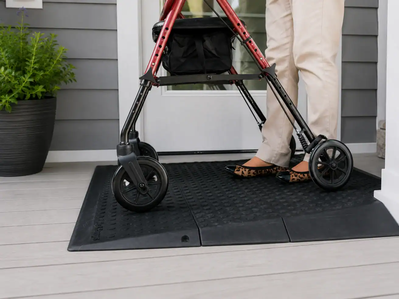 Person using a red walker stands on a textured black rubber mat in front of a white door, wearing beige pants and leopard-print flats. A potted plant is visible to the left.