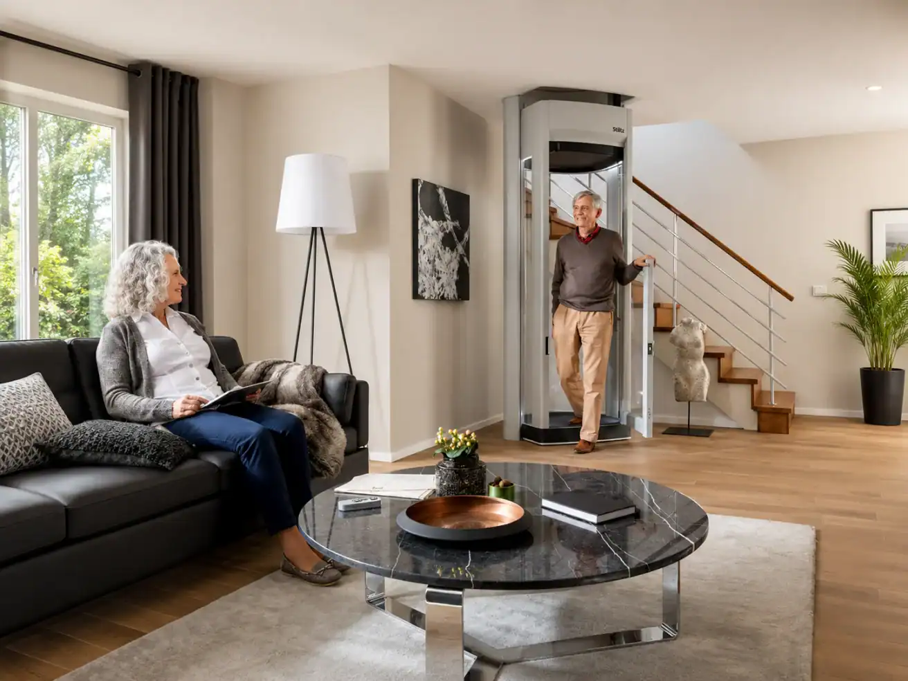An older man exits a residential home elevator as an older woman sits on a sofa reading in a modern living room with a staircase and large windows.