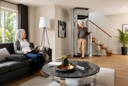 An older man exits a residential home elevator as an older woman sits on a sofa reading in a modern living room with a staircase and large windows.